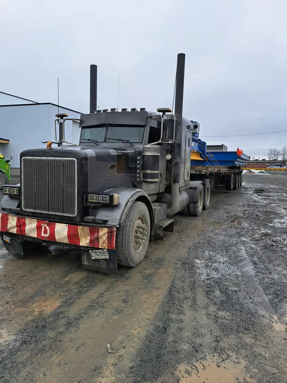 Peterbilt et fardier chargés d’une pièce d’équipement bleue, devant un garage